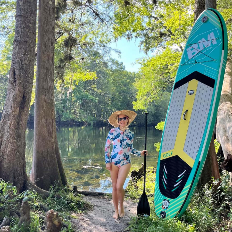 Woman holding a paddle and a green inflatable paddleboard on a forested lake path.