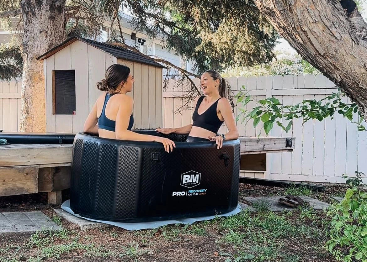 Two women sitting in a portable ice tub outdoors, surrounded by trees and a wooden shed.