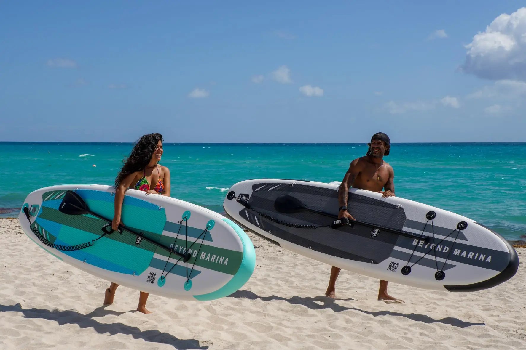 Two people holding inflatable paddle boards on a beach with clear blue water and sky.
