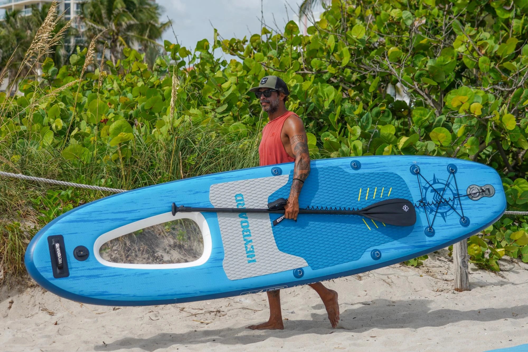 Man holding a blue SUP board on a sandy beach with greenery in the background