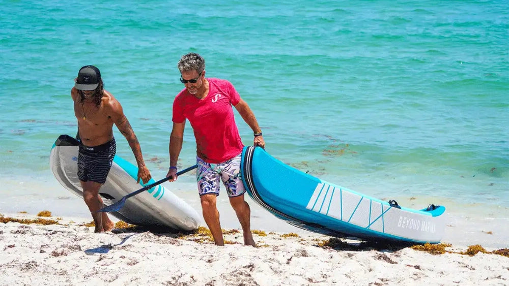 Two people carrying two portable kayaks on a beach with clear blue water in the background